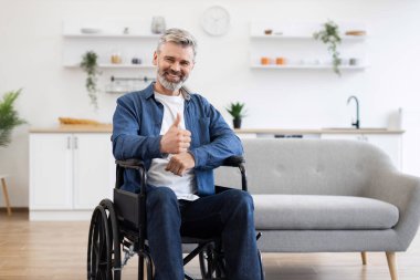 Tekerlekli sandalyede oturan kıdemli beyaz adam içeride pozitifliğini gösteren olumlu bir jest yapıyor. Resim, modern ev ortamında erişilebilirlik, taşınabilirlik yardımı ve neşeli tavırlar sergiliyor.