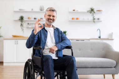 Tekerlekli sandalyedeki son sınıf beyaz erkek gülümsüyor, iyi bir jest yapıyor, modern, parlak bir mutfakta oturuyor. Resim, ev ortamında güven, kapsayıcılık, pozitiflik ve erişilebilirliği tasvir eder.