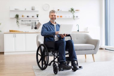 Tekerlekli sandalyede oturan, rahat bir sandalyede oturan, dizüstü bilgisayarda çalışan, üretkenlik ve ev ortamında bağımsız yaşam tarzını sergileyen olgun, beyaz bir adam..