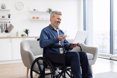 Tekerlekli sandalyede dizüstü bilgisayar kullanan olgun bir beyaz adam parlak bir evde görüntülü konuşma yapıyor. Dizüstü bilgisayar elinde, iletişimle meşgul. Modern iç mekanlarda tekerlekli sandalye erişilebilirliği