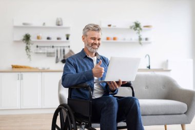 Evde tekerlekli sandalyede oturan yetişkin bir beyaz adam video görüşmesi için dizüstü bilgisayar kullanıyor. Işıl ışıl döşenmiş iç mekan profesyonel ama ulaşılabilir çalışma alanı ortamı yaratıyor