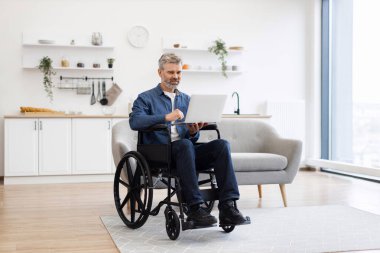 Tekerlekli sandalyedeki olgun beyaz adam evde görüntülü konuşma yapıyor, iletişim kuruyor. Teknoloji, dizüstü bilgisayar kullanan profesyonel veya kişisel bağlantılar için kapsayıcılığı ve etkileşimi teşvik eder.