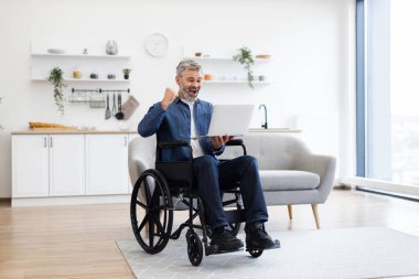 Tekerlekli sandalyedeki yetişkin erkek, zafer için kolunu kaldırıyor, içeride oturuyor, dizüstü bilgisayar kullanırken başarıyı kutluyor, heyecan ve neşe ifade ediyor, hafif iç tasarımla, modern mobilyalarla