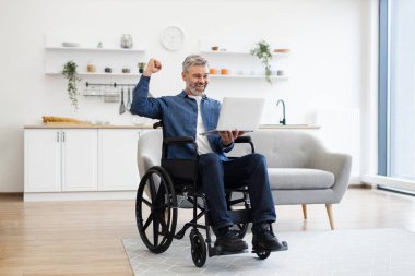 Tekerlekli sandalyede oturan olgun beyaz adam, el kaldırarak başarıyı kutlayan dizüstü bilgisayarını kullanıyor..