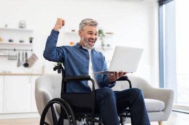 Modern oturma odasında dizüstü bilgisayar kullanırken kutlama için elini kaldıran tekerlekli sandalyedeki olgun beyaz adam. Başarı, pozitiflik ve kapsayıcı ve güçlendirici yaşam tarzındaki uzak başarıyı temsil eder..