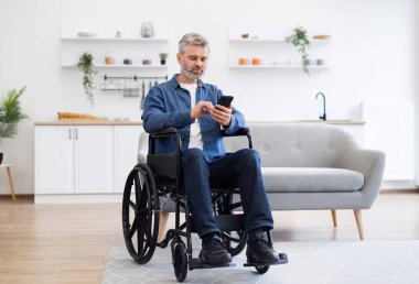 Tekerlekli sandalyede oturan olgun beyaz adam akıllı telefon kullanıyor, modern dekor kullanıyor..