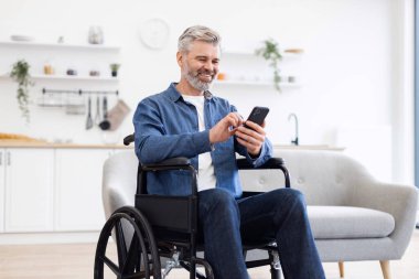Evde tekerlekli sandalyede oturan olgun bir beyaz adam elinde akıllı telefon, etrafı kolaçan ediyor..