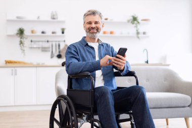 Tekerlekli sandalyede oturan olgun, beyaz bir adam elinde akıllı bir telefon, çağdaş ev içlerinde günlük hayat tarzını gözler önüne seriyor.
