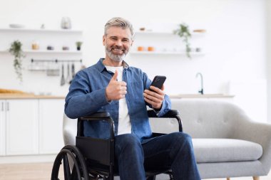 Tekerlekli sandalyede oturan, elinde akıllı telefon tutan, baş parmağını kaldıran, sıradan kıyafetler giyen, modern oturma odasında parlak bir dekorla oturan olgun bir beyaz adam.