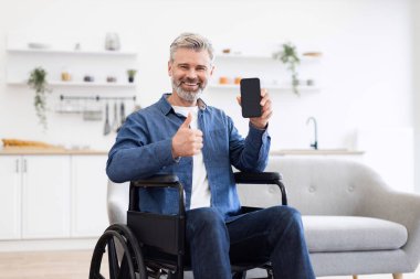Tekerlekli sandalyede beyaz saçlı, akıllı telefon tutan ve modern bir evde gülümseyen olgun bir beyaz adam. Adam onayını başparmak hareketleriyle gösteriyor, bağımsızlığı ve bağlantıyı vurguluyor.