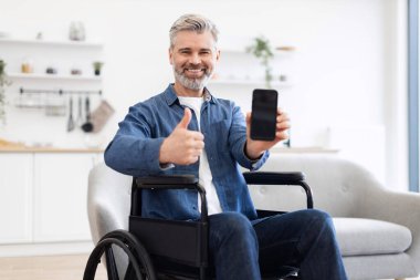 Tekerlekli sandalyede beyaz saçlı, akıllı telefon tutan ve modern bir evde gülümseyen olgun bir beyaz adam. Adam onayını başparmak hareketleriyle gösteriyor, bağımsızlığı ve bağlantıyı vurguluyor.