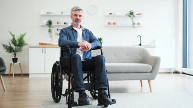 Tekerlekli sandalyede oturan kıdemli beyaz adam içeride pozitifliğini gösteren olumlu bir jest yapıyor. Resim, modern ev ortamında erişilebilirlik, taşınabilirlik yardımı ve neşeli tavırlar sergiliyor.