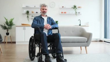 Tekerlekli sandalyedeki son sınıf beyaz erkek gülümsüyor, iyi bir jest yapıyor, modern, parlak bir mutfakta oturuyor. Resim, ev ortamında güven, kapsayıcılık, pozitiflik ve erişilebilirliği tasvir eder.