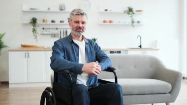 Tekerlekli sandalyede oturan kıdemli beyaz adam içeride pozitifliğini gösteren olumlu bir jest yapıyor. Resim, modern ev ortamında erişilebilirlik, taşınabilirlik yardımı ve neşeli tavırlar sergiliyor.