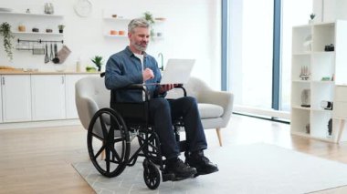 Tekerlekli sandalyede oturan, rahat bir sandalyede oturan, dizüstü bilgisayarda çalışan, üretkenlik ve ev ortamında bağımsız yaşam tarzını sergileyen olgun, beyaz bir adam..