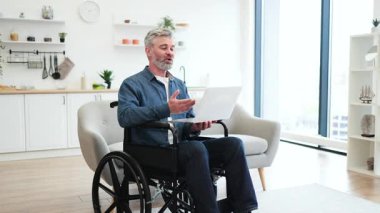 Tekerlekli sandalyede dizüstü bilgisayar kullanan olgun bir beyaz adam parlak bir evde görüntülü konuşma yapıyor. Dizüstü bilgisayar elinde, iletişimle meşgul. Modern iç mekanlarda tekerlekli sandalye erişilebilirliği