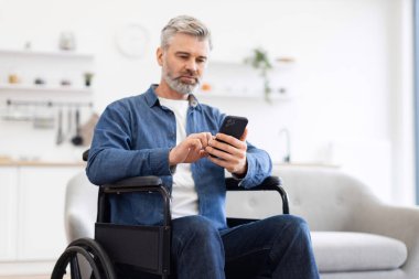 Akıllı telefon kullanarak tekerlekli sandalyede oturan olgun bir beyaz adam. Erişilebilirlik, dijital iletişim ve bağımsızlık odaklı ev düzeni. Teknoloji temalarını destekliyor
