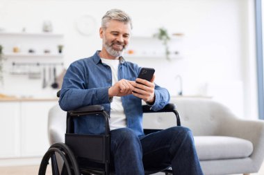 Akıllı telefon kullanarak tekerlekli sandalyede oturan olgun bir beyaz adam. Erişilebilirlik, dijital iletişim ve bağımsızlık odaklı ev düzeni. Teknoloji temalarını destekliyor