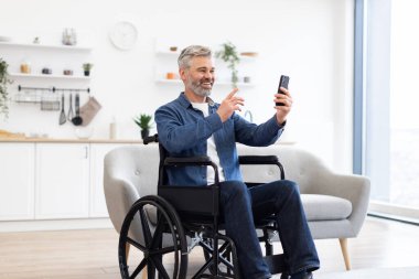 Modern, parlak oturma odasında akıllı telefon kullanarak tekerlekli sandalyedeki olgun beyaz adam. Kapsayıcı teknoloji kullanımı ve bağlanabilirliği gösterir.