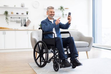 Modern, parlak oturma odasında akıllı telefon kullanarak tekerlekli sandalyedeki olgun beyaz adam. Kapsayıcı teknoloji kullanımı ve bağlanabilirliği gösterir.