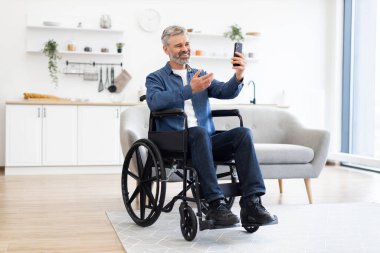 Beyaz saçlı, tekerlekli sandalyede oturan beyaz bir adam video görüşmesi için akıllı telefon kullanıyor. İç dekorasyon sıcaklığı ve modern tasarımı iletir, bağlantı, bağımsızlık ve erişilebilirliği vurgular.