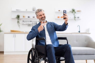 Tekerlekli sandalyedeki olgun beyaz erkek telefonuyla selfie çekiyor, günlük kotlar giyiyor, içeride.