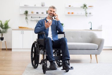 Tekerlekli sandalyede oturan olgun beyaz adam ev ortamında cep telefonuyla konuşuyor. Gündelik yaşam anı parlak ve modern bir iç dekorla sunuluyor..