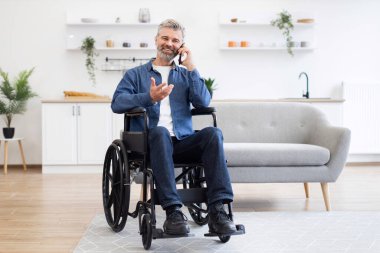 Tekerlekli sandalyede oturan olgun beyaz adam ev ortamında cep telefonuyla konuşuyor. Gündelik yaşam anı parlak ve modern bir iç dekorla sunuluyor..