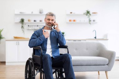 Olgun, beyaz, sakallı bir adam. Parlak, modern oturma odasında tekerlekli sandalyede oturuyor. İletişim için cep telefonunu kullanıyor.
