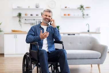 Olgun, beyaz, sakallı bir adam. Parlak, modern oturma odasında tekerlekli sandalyede oturuyor. İletişim için cep telefonunu kullanıyor.