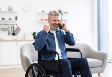 Tekerlekli sandalyede oturan, akıllı telefondan konuşan, modern oturma odasında yumruklarını sıkarak heyecanını ifade eden olgun beyaz bir adam.