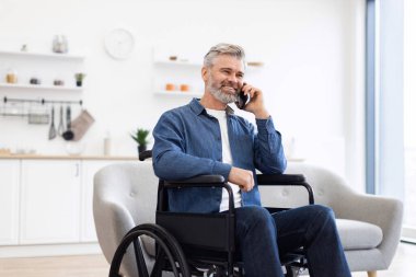 Tekerlekli sandalyede oturan, telefon kullanan, kot giyen, kapalı alanda gülümseyen, mutluluk ve bağımsızlığını ifade eden olgun, beyaz bir adam.