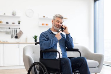 Tekerlekli sandalyede oturan, telefon kullanan, kot giyen, kapalı alanda gülümseyen, mutluluk ve bağımsızlığını ifade eden olgun, beyaz bir adam.