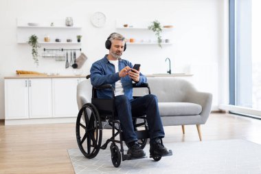 Tekerlekli sandalyedeki olgun beyaz adam, modern yaşam alanında kulaklık ve telefon kullanarak müzik dinlemekten hoşlanıyor. Müzik, bağımsızlık ve teknolojinin kullanımına olan tutkusunu ifade eder.