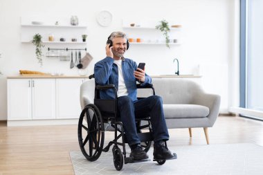 Akıllı telefon kullanan, müzik dinleyen ve parlak oturma odasında gülümseyen, tekerlekli sandalyedeki olgun beyaz adam. Kulaklıklı sıradan kıyafetler giymek, evde rahatlama ve mutluluğun tadını çıkarmak..