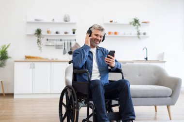 Akıllı telefon kullanan, müzik dinleyen ve parlak oturma odasında gülümseyen, tekerlekli sandalyedeki olgun beyaz adam. Kulaklıklı sıradan kıyafetler giymek, evde rahatlama ve mutluluğun tadını çıkarmak..