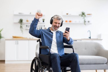 Tekerlekli sandalyedeki olgun beyaz adam telefonu kullanıyor, kulaklıkla müzik dinliyor, gülümsüyor ve kutluyor. İçi rahat modern ev ortamları. Neşe müziği ve erişilebilirlik artışı kavramı.