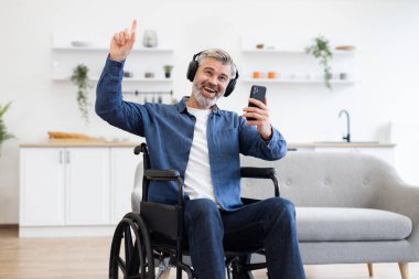 Tekerlekli sandalyedeki olgun beyaz adam modern iç mekanlarda telefonla kulaklıkla müziğin keyfini çıkarıyor. Neşeyi, kapsayıcılığı ve melodiyi kucaklayan aşk. Pozitifliği ve kendini ifade etmeyi vurgula.