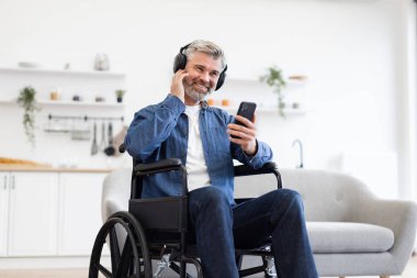 Tekerlekli sandalyedeki olgun beyaz adam kulaklık takıyor ve telefon kullanıyor. Parlak oturma odası ortamında neşeli ruh hali, bağımsızlık ve pozitiflik sergiliyor..