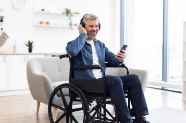 Tekerlekli sandalyedeki olgun beyaz adam kulaklık takıyor ve telefon kullanıyor. Parlak oturma odası ortamında neşeli ruh hali, bağımsızlık ve pozitiflik sergiliyor..