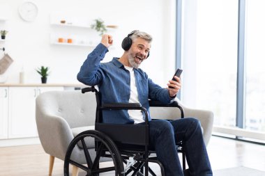 Olgun, beyaz bir adam, yaklaşık 50 'li yaşlarda, tekerlekli sandalyede oturuyor, akıllı telefonuyla meşgul, kulaklık takıyor, hevesle gülümsüyor, müzikten hoşlanıyor, mutluluğunu ifade ediyor.
