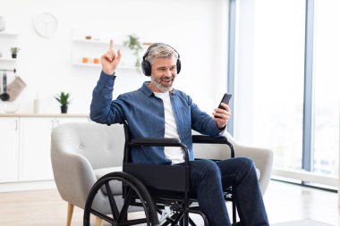 Olgun, beyaz bir adam, yaklaşık 50 'li yaşlarda, tekerlekli sandalyede oturuyor, akıllı telefonuyla meşgul, kulaklık takıyor, hevesle gülümsüyor, müzikten hoşlanıyor, mutluluğunu ifade ediyor.
