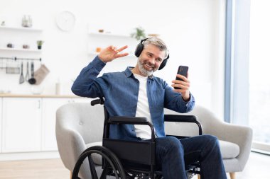 Tekerlekli sandalyede oturan, akıllı telefon ve kulaklık kullanırken gülümseyen, müzikten hoşlanan olgun beyaz adam..