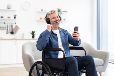 Tekerlekli sandalyede akıllı telefon kullanan, kulaklıkla müzik dinleyen, neşeli ve rahat ev ortamında ritimle hareket eden olgun bir erkek..