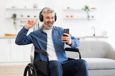 Tekerlekli sandalyedeki olgun beyaz adam kulaklık takıyor ve mutfak ortamında mutlu bir şekilde dans ediyor.