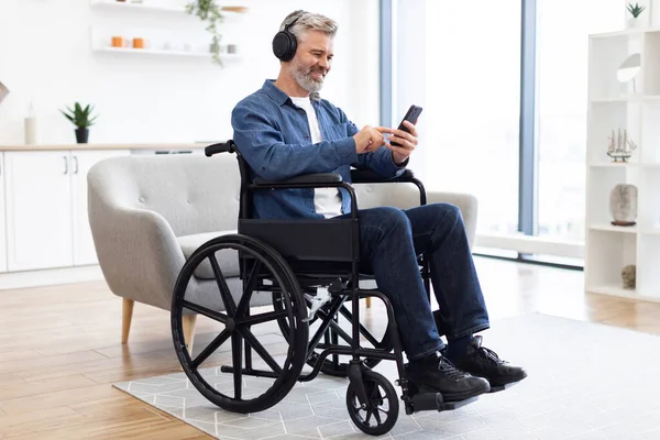 Tekerlekli sandalyedeki olgun beyaz adam kulaklıkla müzik dinlemek için akıllı telefon kullanıyor. İçeride gülümsüyor ve rahatlıyor. Doğal ışık ve şık mobilyaları olan rahat bir modern ev ortamı..