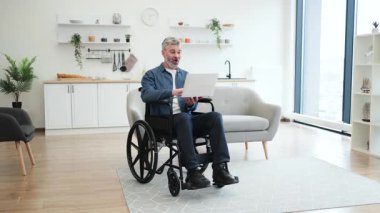 Tekerlekli sandalyedeki yetişkin erkek, zafer için kolunu kaldırıyor, içeride oturuyor, dizüstü bilgisayar kullanırken başarıyı kutluyor, heyecan ve neşe ifade ediyor, hafif iç tasarımla, modern mobilyalarla