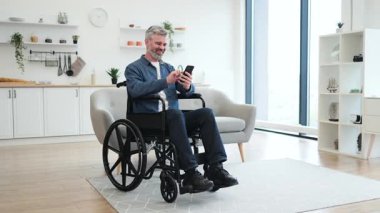 Tekerlekli sandalyede oturan olgun beyaz adam akıllı telefon kullanıyor, modern dekor kullanıyor..