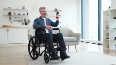 Modern, parlak oturma odasında akıllı telefon kullanarak tekerlekli sandalyedeki olgun beyaz adam. Kapsayıcı teknoloji kullanımı ve bağlanabilirliği gösterir.