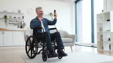 Beyaz saçlı, tekerlekli sandalyede oturan beyaz bir adam video görüşmesi için akıllı telefon kullanıyor. İç dekorasyon sıcaklığı ve modern tasarımı iletir, bağlantı, bağımsızlık ve erişilebilirliği vurgular.